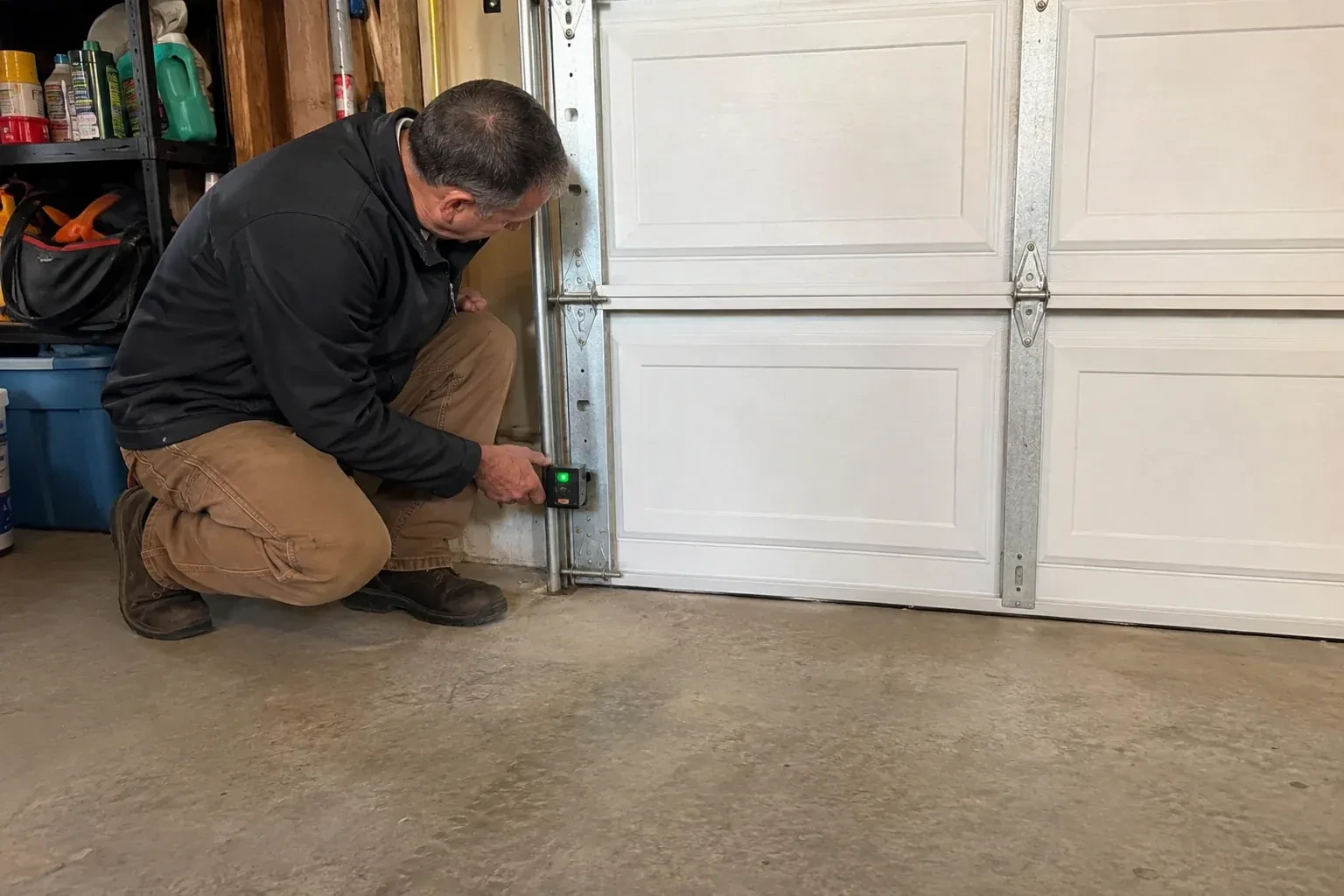 Technician kneeling beside a white paneled garage door, holding a small black box with a green light against the vertical track. — Photo-Eye Sensor Alignment or Replacement, Burlington
